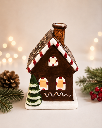 A ceramic gingerbread house ornament with a lit window, decorated with red and white icing details, a small green tree, and a brown chimney, set against a warm background with bokeh lights and holiday greenery.