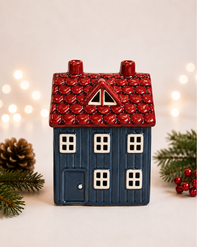 A ceramic Christmas house candle holder with a dark blue body and a textured red roof, styled with pine branches, a pinecone, and warm bokeh fairy lights in the background.