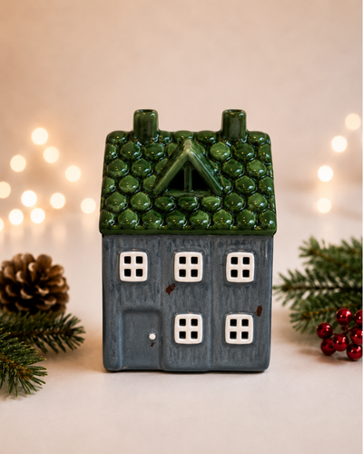 A small ceramic house-shaped candle holder or decorative figurine with a textured green roof, dark blue-gray body, and white window panes, styled with pine branches, a pinecone, and red berries against a warm blurred background with string lights.