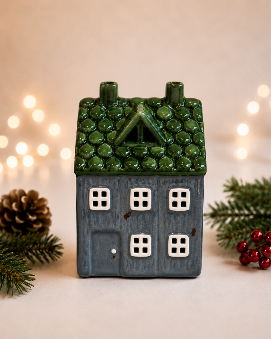 A small ceramic house-shaped candle holder or decorative figurine with a textured green roof, dark blue-gray body, and white window panes, styled with pine branches, a pinecone, and red berries against a warm blurred background with string lights.