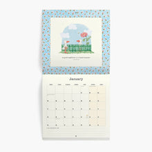 A hanging monthly desk calendar shown open against a light gray background. The top page features a pastel illustration of a garden scene with a pink flamingo, green fence, and blue sky, bordered by a light blue patterned frame.